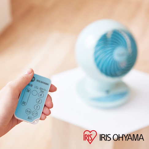 手持藍色遙控器，上面印有「IRIS OHYAMA」字樣及多個按鈕，包含風量強弱、擺頭及定時功能。背景為淺藍色圓形空氣循環扇，扇葉為藍色螺旋狀。