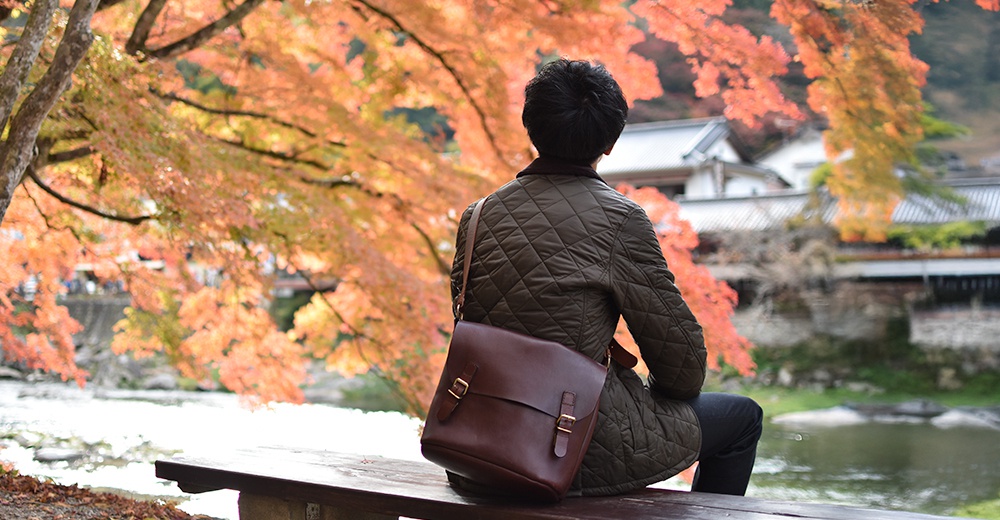 在秋楓背景下，一位男士背對鏡頭，坐在長凳上，肩上掛著一個褐色的皮革郵差包。