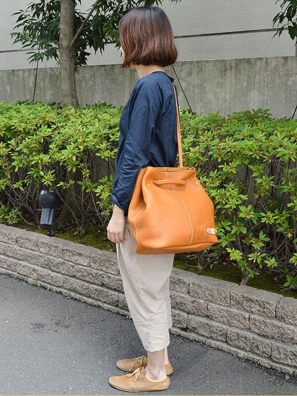 一位女士身穿深藍色上衣和米色褲子，肩上斜背著一個橘棕色皮革托特包。包包有抽繩閉合設計，肩帶細長，整體為圓潤的桶狀造型。