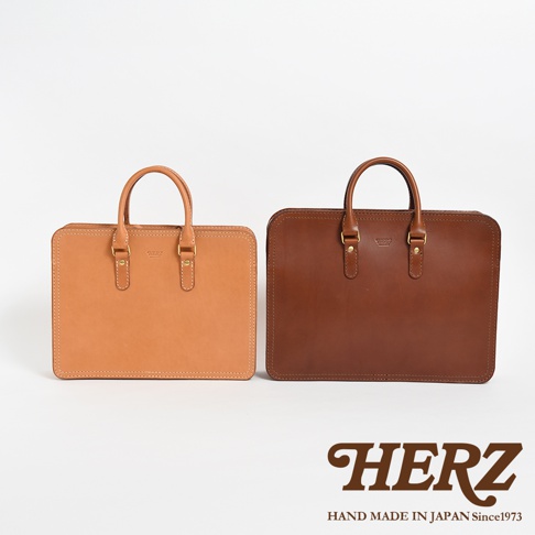 兩個職人手作皮革手提商務公事包，分別為淺棕色和深棕色，均為簡約長方形設計，配有雙提把與金色金屬扣件。