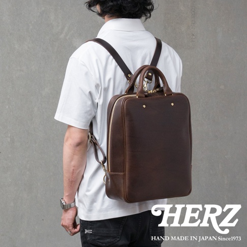 一名男子身著白色T恤和黑色牛仔褲，背著一個深棕色皮革後背包，包包上有金色拉鍊和細節。