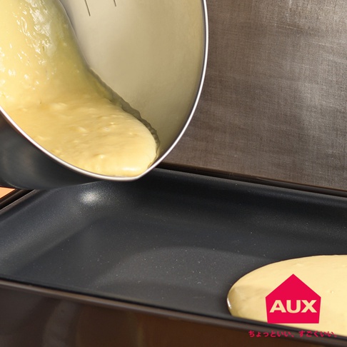 銀色不鏽鋼刻度攪拌鍋傾倒金黃色麵糊，部分麵糊落在黑色平底烤盤上。