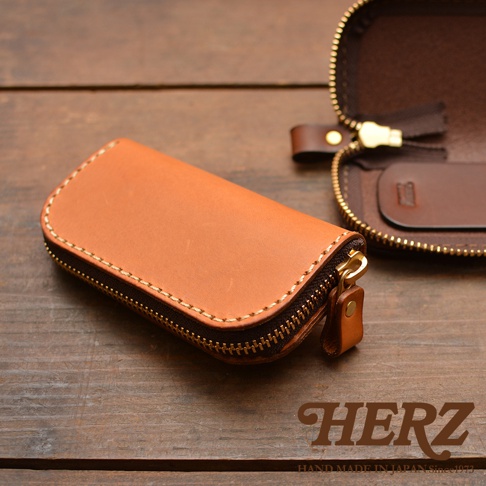 一個棕褐色皮革橢圓形拉鍊鑰匙包，帶有金屬拉鍊和四個掛鉤，位於深色木質背景上，旁邊展示著一個打開的同款鑰匙包。