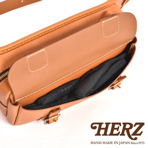 淺棕色皮革斜背包，帶有白色縫線和黃銅色金屬扣。包蓋打開，露出內部裝有黑色手套的隔層。