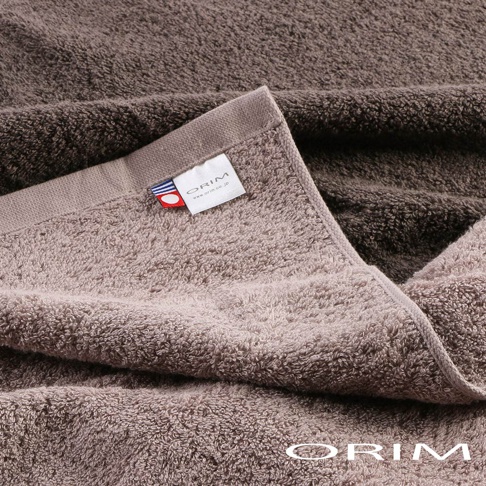 兩條深棕色和淺棕色的今治毛巾，柔軟蓬鬆的毛圈材質，其中一條毛巾的邊緣縫有印有 ORIM 品牌標誌和網址的白色標籤，標籤旁還有藍、白、紅三色條紋裝飾。