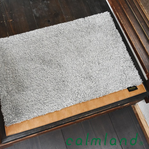 Ward Robe M 尺寸的灰色長絨毛玄關地墊，邊緣飾有棕色仿麂皮飾帶，置於木質地板上。