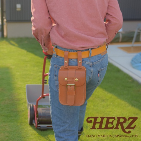 一個棕色皮革腰袋，帶有金屬扣和品牌標誌，掛在穿著牛仔褲和紅色襯衫的人的腰部。