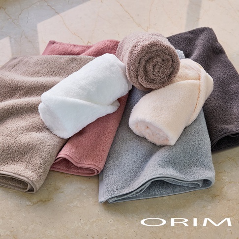 多條不同顏色的 ORIM 日本今治認證毛巾堆疊在一起，包含白色、粉色、米色、棕色、灰色等，毛巾材質呈現柔軟厚實的觸感，整體畫面展示了精緻的布料質感與豐富的色系選擇。