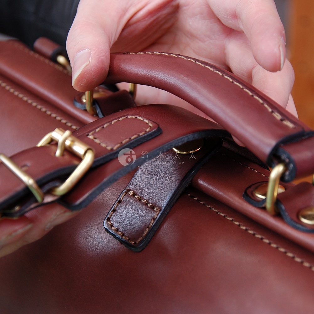 手持棕色皮革杜勒斯包的提把，展示包包的細節，包括縫線、黃銅扣環和皮革材質。