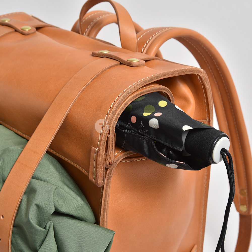 棕色皮革後背包，一側有綠色衣物，另一側插著一把黑色傘，傘面有彩色圓點圖案。