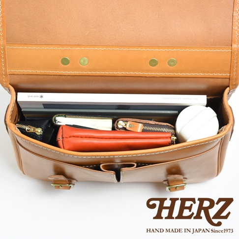 棕色皮革製成的手提包，內部空間擺放著書籍、筆記本、橘色皮夾、黑色皮夾和白色圓形物品。包包外觀有金屬釦和縫線細節。