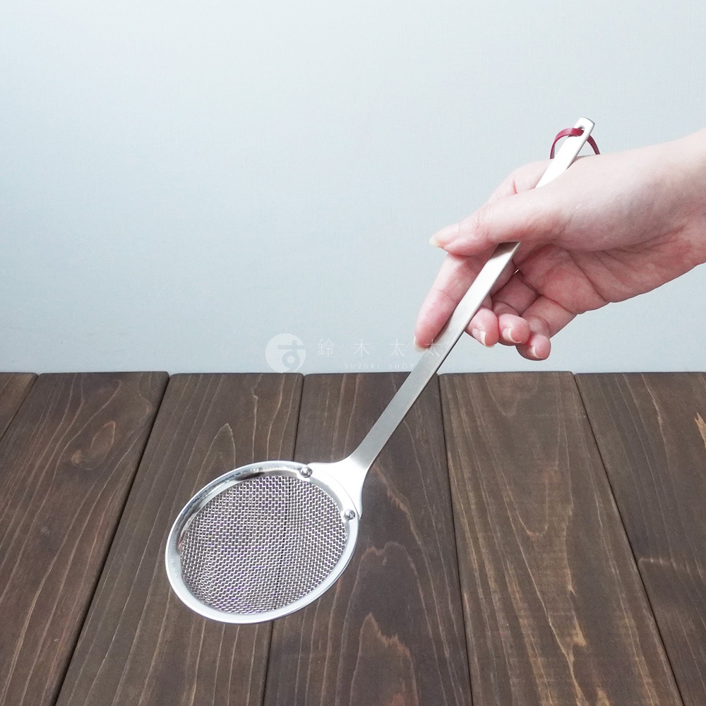 一只手握著不鏽鋼萬用撈油勺，勺頭為橢圓形網篩，細密網孔，勺柄為銀色金屬材質，末端有紅色掛繩。