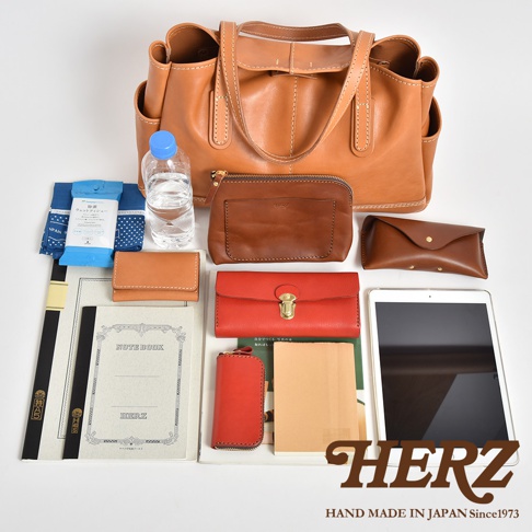 俯視視角下展示日本HERZ職人手作皮革托特包，包包位於上方，周圍整齊擺放各類日常用品，包括筆記型電腦或平板、筆記本、紅色與棕色長夾、眼鏡盒、水瓶、濕紙巾與小物包，整體呈現溫潤的皮革質感與日系精緻風格。