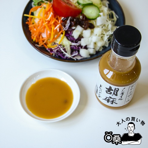 玻璃瓶裝的無油芝麻沙拉醬，瓶身有日文標籤，醬汁呈現淺棕色。旁邊有一個白色小碟，裝有少量相同顏色的沙拉醬。背景是裝有豐富蔬菜的沙拉。