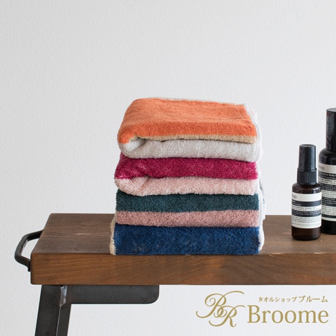 一疊由今治純棉製成的毛巾，呈現橘色、米色、桃紅、淺粉、深綠、灰粉、深藍等色塊混色，質感柔軟，擺放在木質層架上。