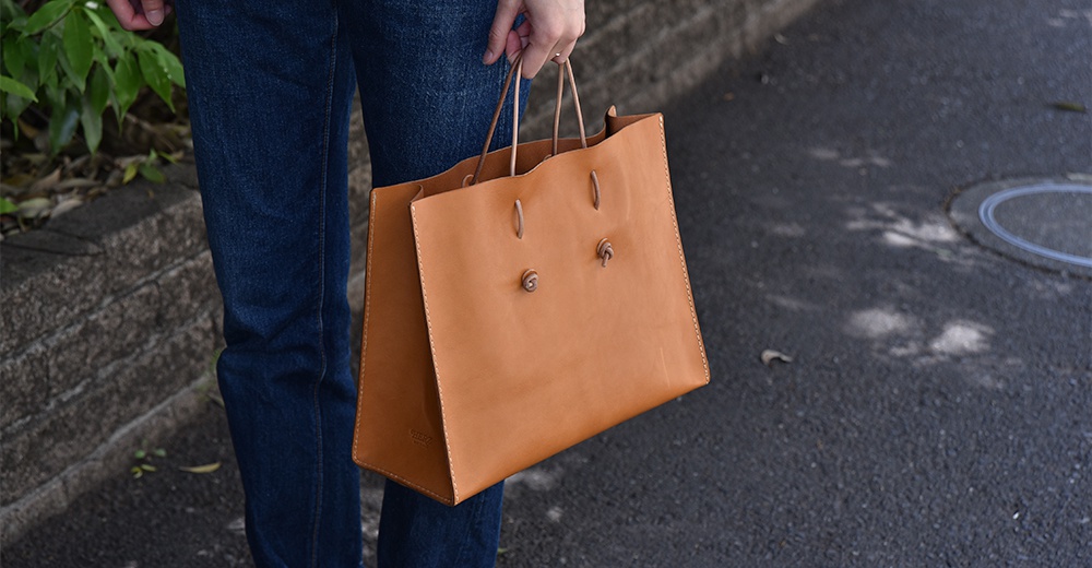 職人手作皮革提袋，淺棕色，採用光滑皮革材質，設計簡約，提把為細長皮革繩，袋面有兩處皮革打結裝飾，整體呈現自然質感。