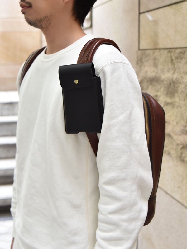 黑色皮革三用手機收納包，附有金色按扣，固定在白色長袖上衣的右肩帶上，搭配棕色皮革背包。