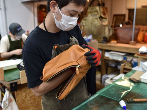 一位戴著口罩和手套的工匠正在檢查一個 tan 色皮革立體運動斜背包，包包有銀色拉鍊，背景是工作室。