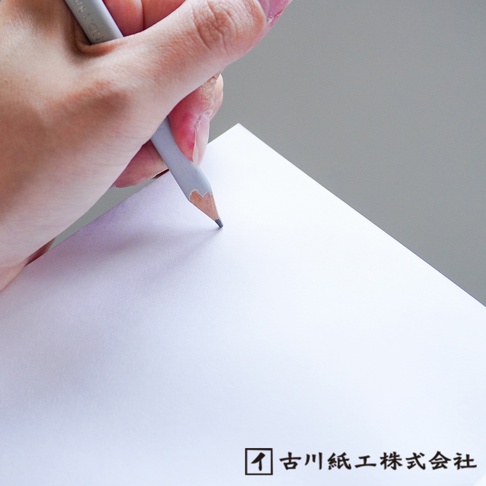 手握淺灰色鉛筆，在純白色紙張上書寫。紙張質感細緻，角落有「古川紙工株式會社」黑色方形標誌。