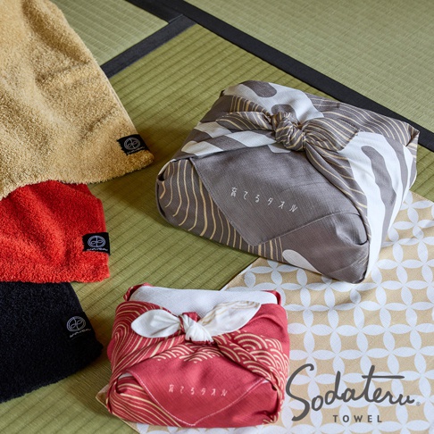 綠色榻榻米墊上擺放著三條不同顏色的毛巾，其中一條黃色，一條紅色，一條黑色，每條毛巾的右下角都有黑色標籤。旁邊是兩件用花紋布包裹的禮品，一件用灰色印有白色和金色線條的花紋布包裹，另一件用紅色印有金色圓紋的花紋布包裹。禮品的包裹方式是日式風呂敷，繫著蝴蝶結。在圖片的右下角有「Sodateru TOWEL」的品牌標誌。