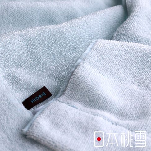淺藍色異素材厚手迷你浴巾，質地柔軟，帶有明顯的織紋。左側有黑色標籤，上面印有「HIORIE」字樣。右下角有日文「日本桃雪」字樣及紅色圓點標誌。