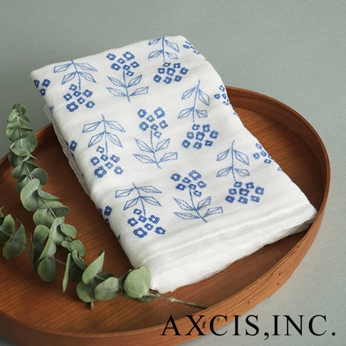 白色純棉花草紗布大拭巾，印有藍色花草圖案，質地輕柔，置於木質圓盤中，旁有一枝綠葉裝飾。