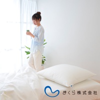 寬大蓬鬆的白色方枕，枕套材質柔軟，放在純白色床單上，旁邊有綠葉植物，一位女士手持杯子站在窗邊。