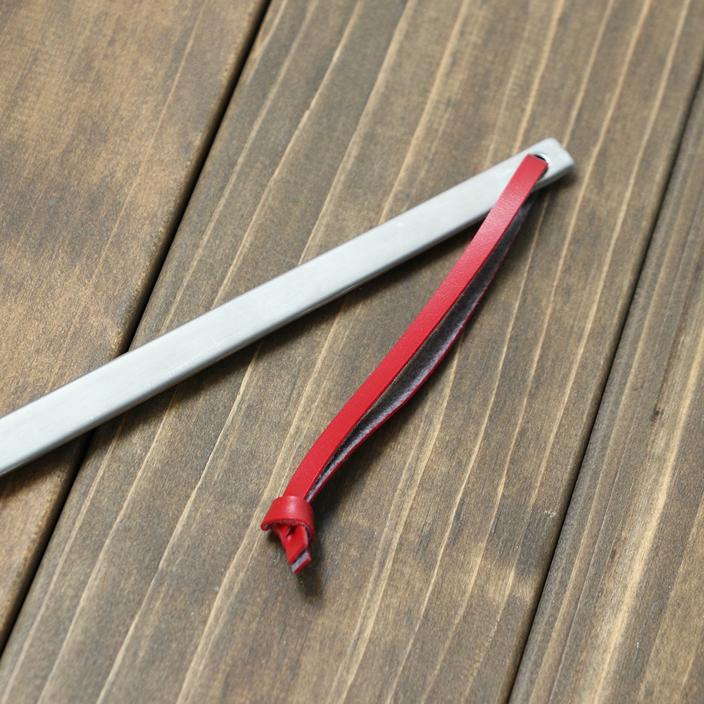 不鏽鋼萬用鍋鏟，鍋鏟主體為銀灰色金屬，扁平設計，末端有一個小孔。一條鮮紅色皮革掛繩穿過孔洞，皮革掛繩末端打結。