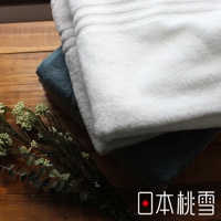 三條今治飯店浴巾堆疊，分別為白色、淺灰色和深藍色。浴巾材質為柔軟的棉，表面有細緻的條紋織法。左側擺放著一束乾燥的綠色植物作為裝飾，背景為木紋桌面。