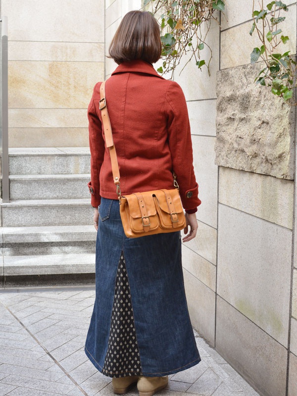 一位女士身穿紅色短外套與牛仔長裙，斜挎一個棕色皮革雙口袋肩背包，包上有金色金屬扣環。