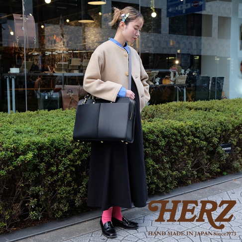 一位女性手提黑色皮革雙口袋兩用商務公事包，包包有拉鍊開合，底部有金色鉚釘裝飾。