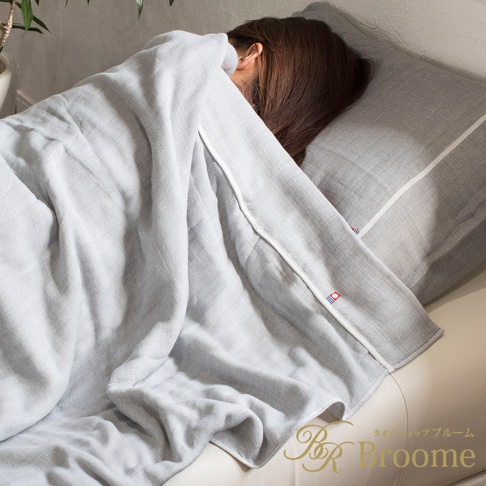 一條淺灰色今治五層紗四季被，覆蓋在一名女士身上，女士頭部枕在同色系的枕頭上，被子邊緣有白色滾邊及藍紅小標誌。