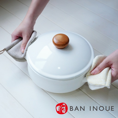 雙手各持一個Tricot棉麻防燙鍋套，分別套在白色鍋子的兩側把手上。鍋套為灰色和米白色，帶有細膩的織紋，鍋蓋中央有一個木質提手。