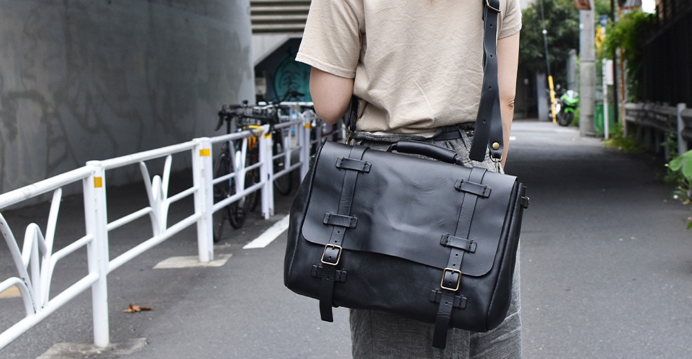 黑色皮革復古郵差包，配有可調節的肩帶和搭扣閉合設計，風格簡潔實用。
