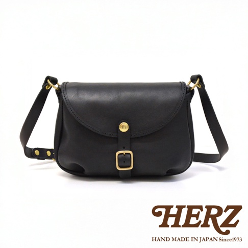 HERZ 品牌黑色皮革斜背包，正面有金屬釦和皮帶裝飾，肩帶可調整，HERZ 手作於日本。