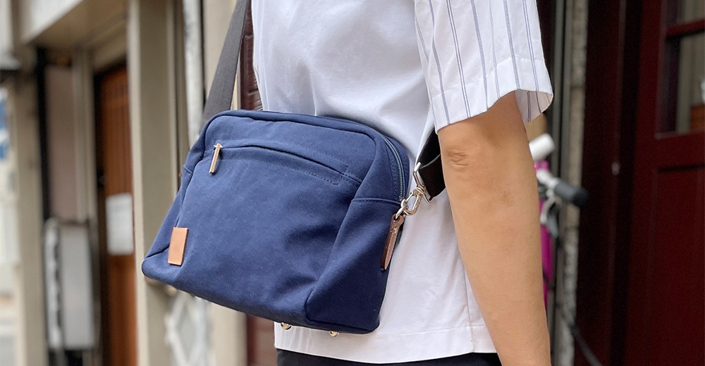 一個斜挎著深藍色石洗帆布旅行側背包的男子，背包上有一個方形的淺棕色皮革標籤。