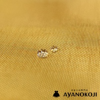 一塊金黃色布料特寫，上面有兩顆圓潤的水珠，右下角有 AYANOKOJI 的 Logo 和日文文字。