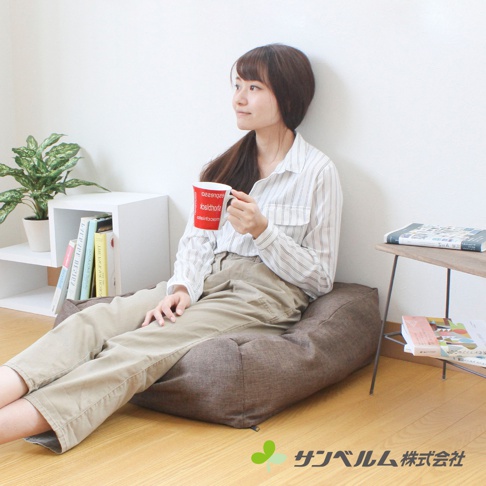 一個女性坐在深棕色、紋理織物的懶骨頭沙發上，她拿著一個紅色的馬克杯，穿著條紋襯衫和卡其褲。