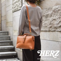 職人手作皮革迷你軟皮杜勒斯包，採用淺棕色皮革製成，配有可調節的長肩帶和頂部提把，包身帶有金屬扣環裝飾。