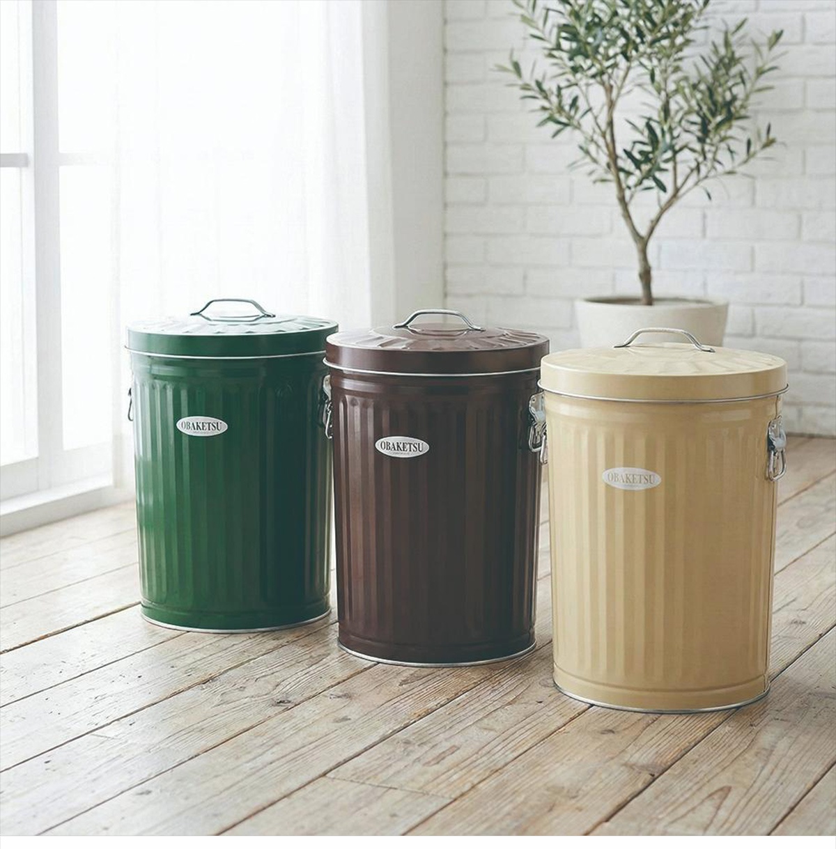 OBAKETSU 品牌三款不同顏色（綠色、棕色、米黃色）的金屬垃圾桶，帶有提把和蓋子，擺放在木質地板上。