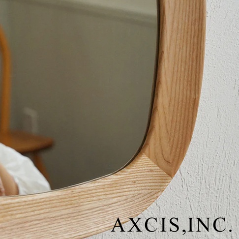 鏡子邊緣為圓潤彎曲的淺色木框，木紋清晰可見，框角處以斜接方式拼接。鏡面反射出室內環境，包含木質椅子和白色衣物。