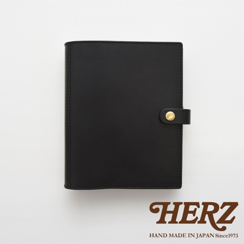 黑色皮革A5手帳/記事本書套，附有金色金屬按鈕和皮革束帶，邊緣縫線整齊，展現職人手作質感。