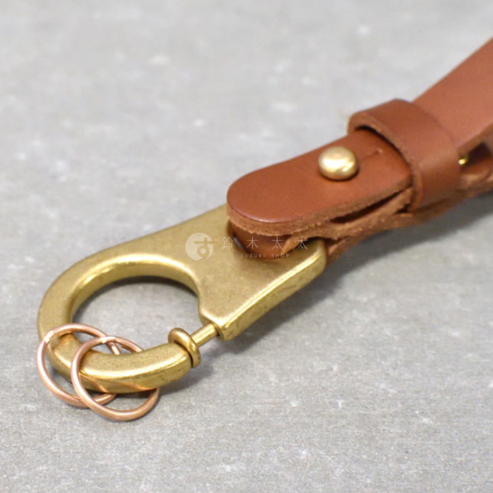 職人手作皮革鑰匙圈，採用棕色皮革搭配黃銅色金屬掛鉤與圓環，皮革以鉚釘固定，質感樸實。