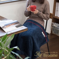 深藍色羊毛感膝蓋毯，邊緣有紅色滾邊，一女子手持紅色杯子，坐在椅子上，膝蓋蓋著毯子，左側為木桌，桌上有打開的雜誌。