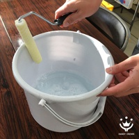 一手持帶有金屬支架的黃色滾筒，將其浸入裝有淺藍色泡沫水的白色塑膠桶中。