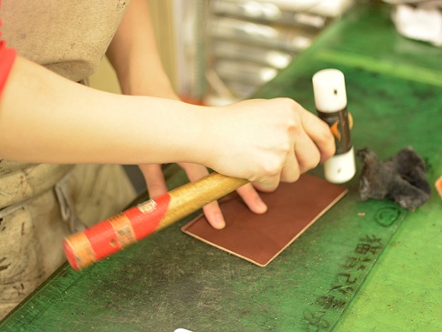 職人手作皮革存摺文件收納夾 (KZ-144)，使用棕色皮革製作，由師傅手持木柄鐵鎚和圓形壓具，在綠色工作台上進行加工。