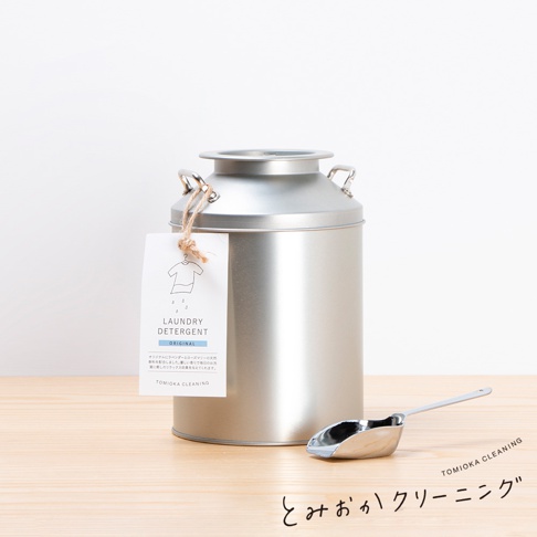 銀色金屬牛奶罐造型洗衣粉罐，附有吊牌，旁邊放著一個銀色金屬量勺。