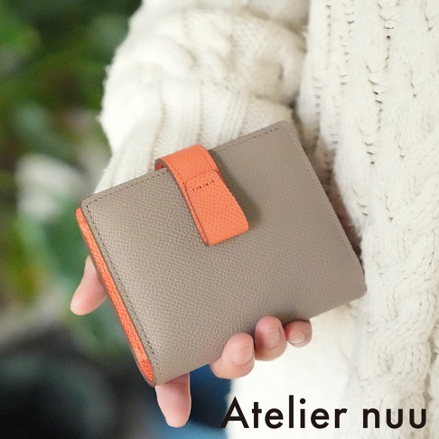 Atelier nuu 品牌 logo 旁，以手持的淺褐色皮革雙折短夾，搭配亮橘色內襯與皮帶扣，展現其精緻的質感。
