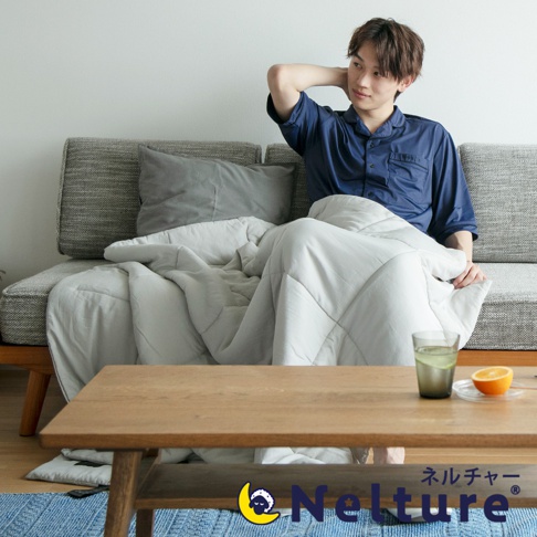 淺灰色 Nemulia 五層散熱涼感單人被，表面帶有菱形格紋縫線，質地輕柔。一位年輕男性身著深藍色睡衣，坐在沙發上，蓋著這件被子，手搭在脖子後方。