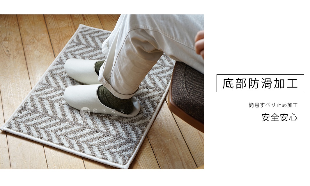 一塊淺棕色與米白色相間，帶有魚骨紋路的短絨毛地墊，鋪設在木質地板上，上面有一雙白色室內拖鞋，露出綠色襪子。